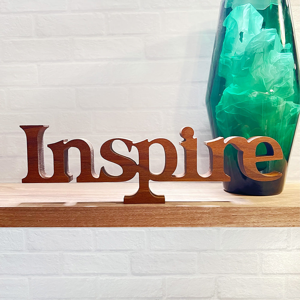 A wooden sign with the standing word - INSPIRE sitting on top of a shelf.